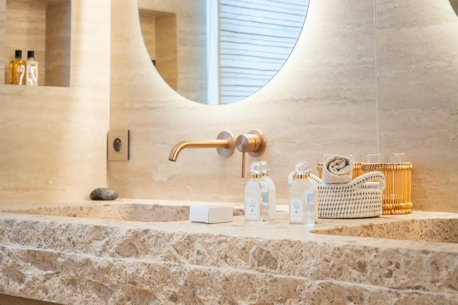 Lavabo de baño moderno con encimera de mármol, espejo redondo, grifo dorado y artículos de aseo en una cesta tejida.
