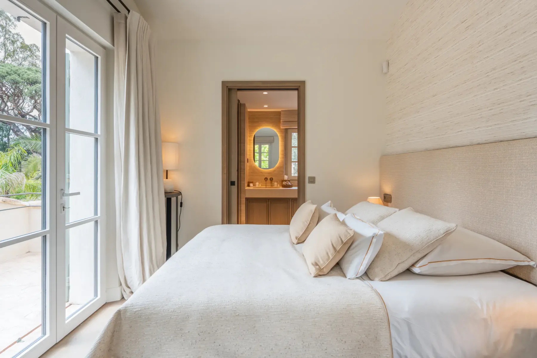 Dormitorio minimalista con una cama acogedora, iluminación suave y una puerta abierta que da a un baño. Un gran ventanal aporta luz natural.