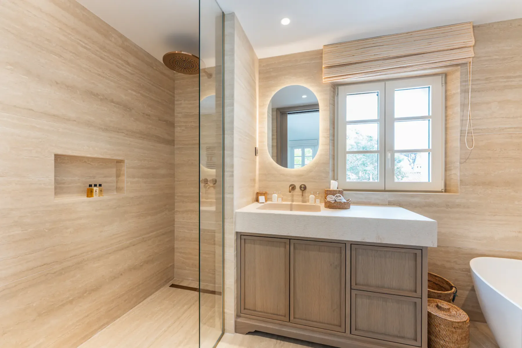 Baño moderno con detalles de madera, un espejo redondo, un gran ventanal, una ducha acristalada y un estante integrado en la pared. La iluminación cálida realza el ambiente acogedor.
