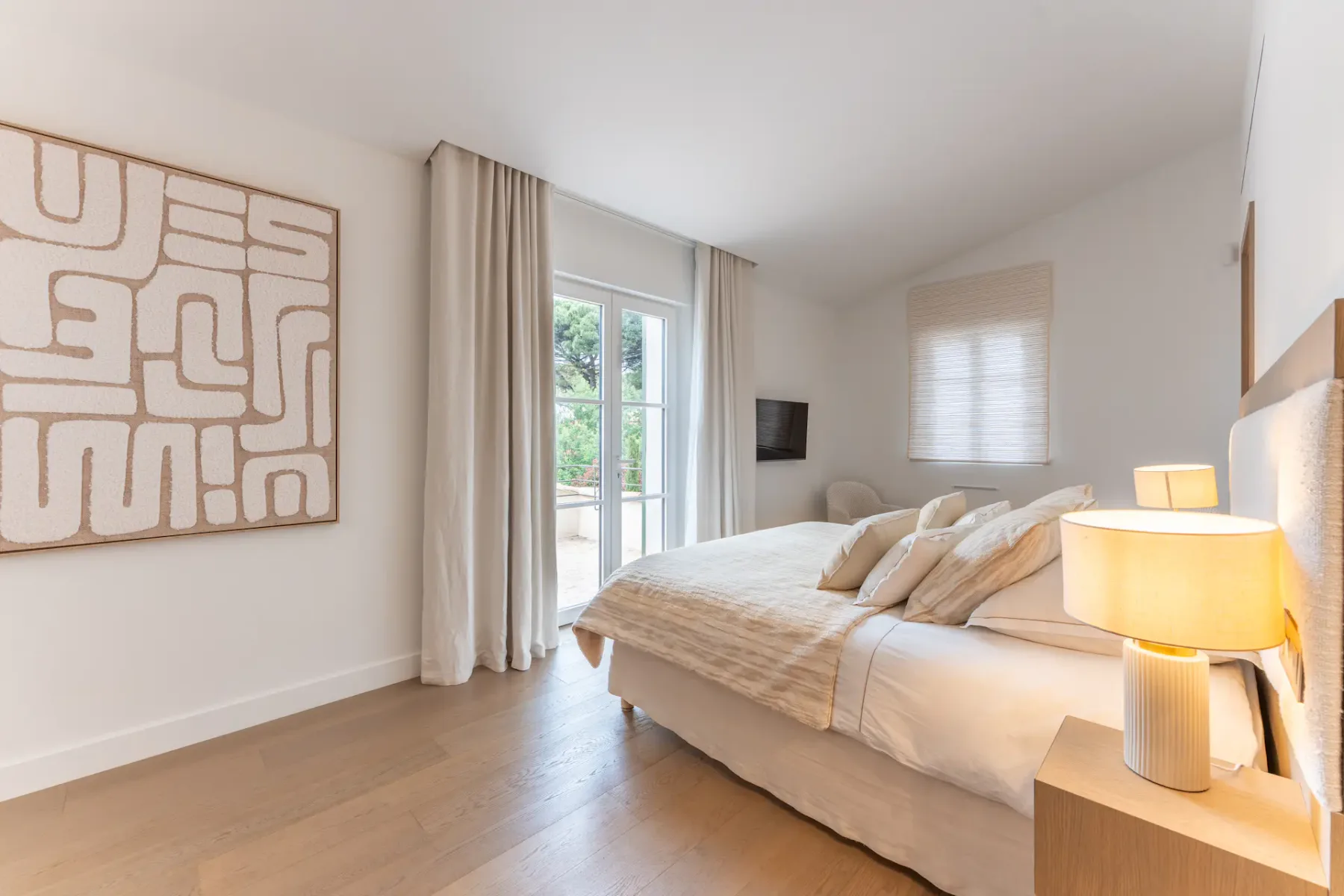 Dormitorio moderno con una cama grande, arte abstracto en la pared, una lámpara de noche y puertas de cristal que dan a un balcón con vegetación en el exterior.
