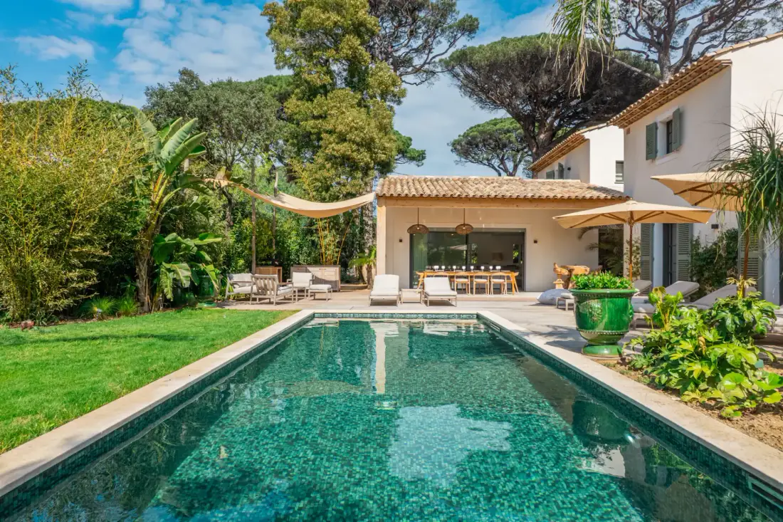 Una piscina está junto a un patio con asientos al aire libre, sombrillas y vegetación.