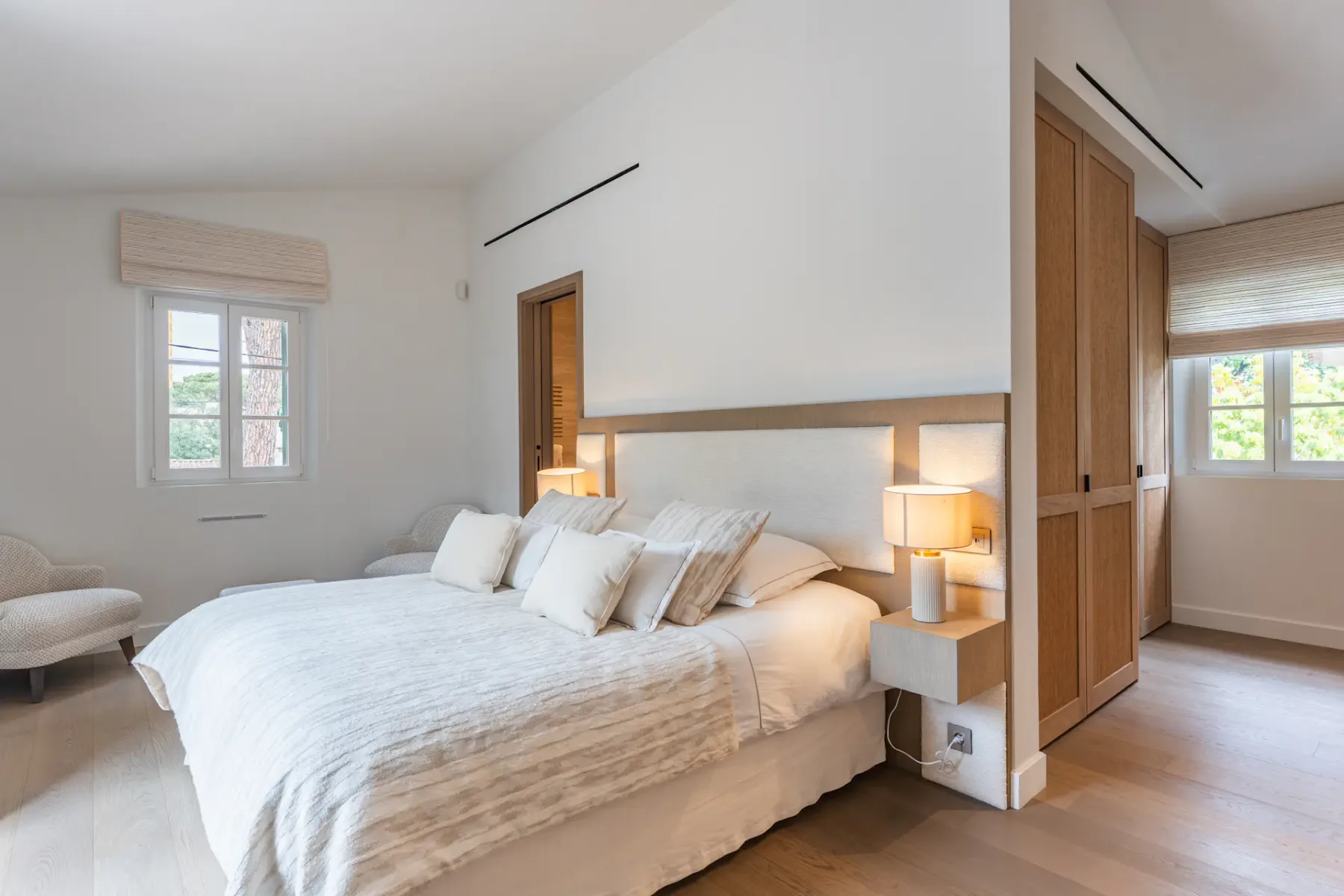 Dormitorio luminoso con una cama grande, ropa de cama blanca, lámparas de noche, un armario de madera y dos ventanas que dejan entrar luz natural.