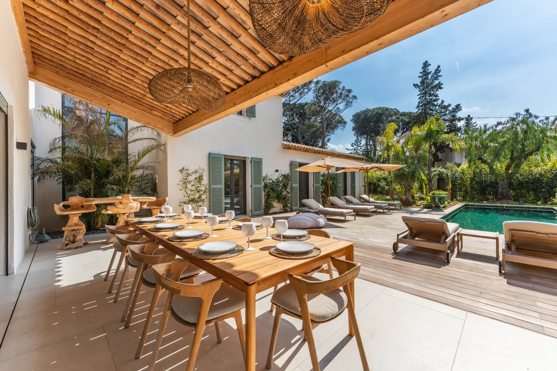 Comedor al aire libre con una larga mesa de madera preparada para una comida, con vistas a una piscina rodeada de tumbonas y plantas tropicales bajo un cielo azul.