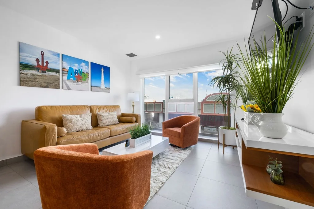 Sink into plush seating and feel the coastal breeze through your private balcony doors. Sunlight fills your airy living space, where vibrant local art and lush greenery invite you to unwind in modern comfort.