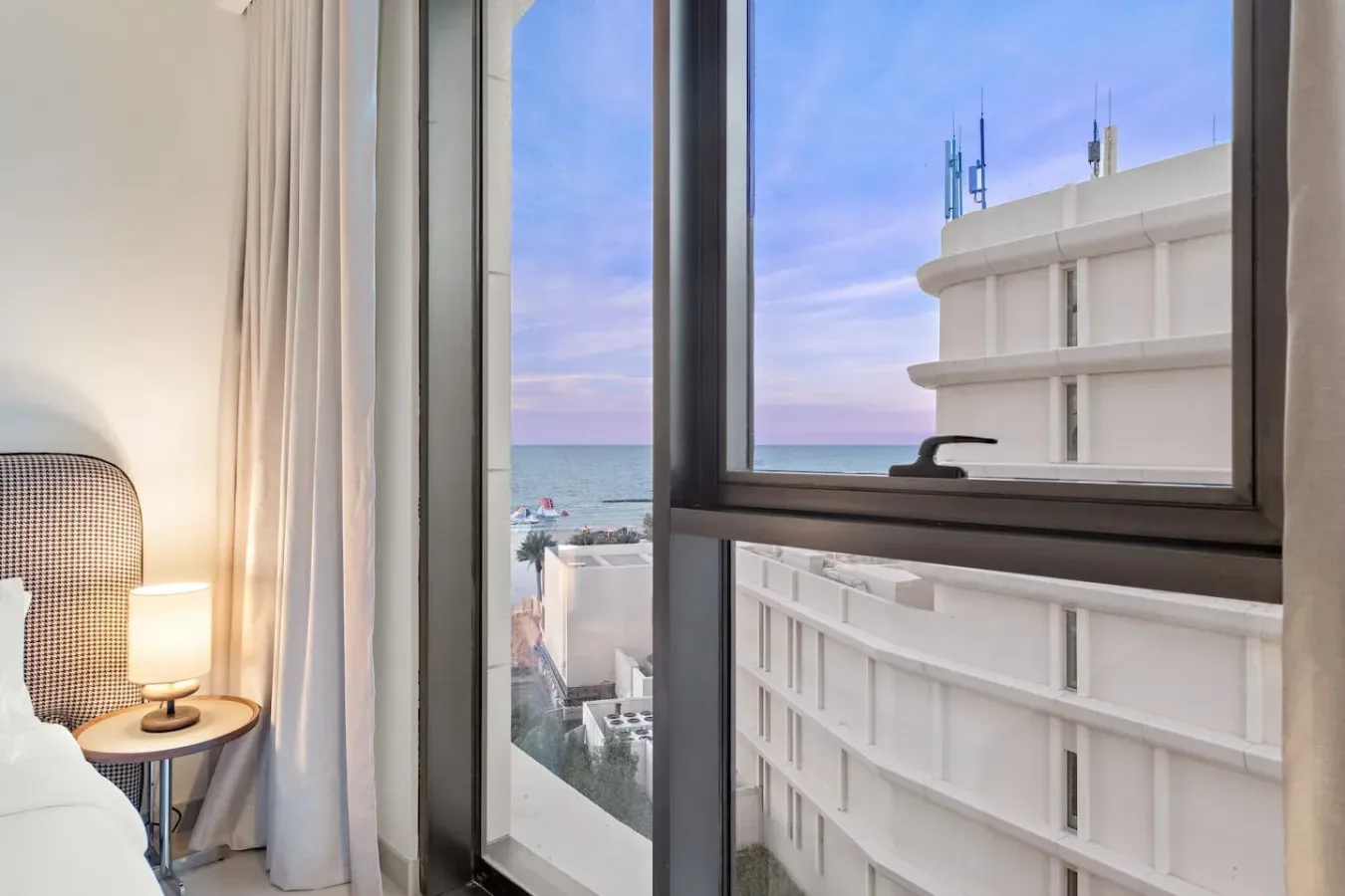 Una ventana de dormitorio da al mar y a un edificio blanco cercano.
