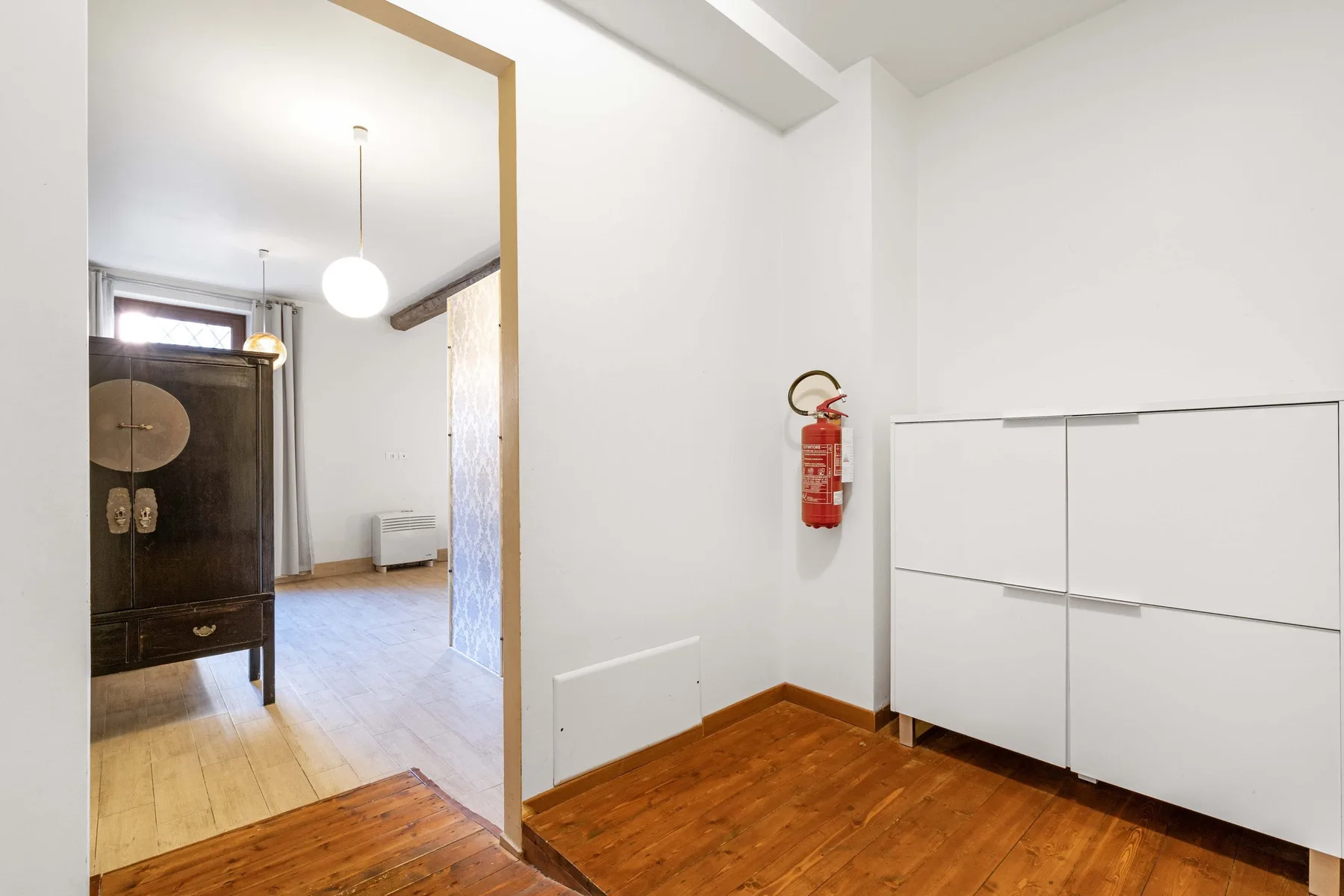 Una habitación luminosa con suelo de madera, armarios blancos y un mueble oscuro en el espacio contiguo.
