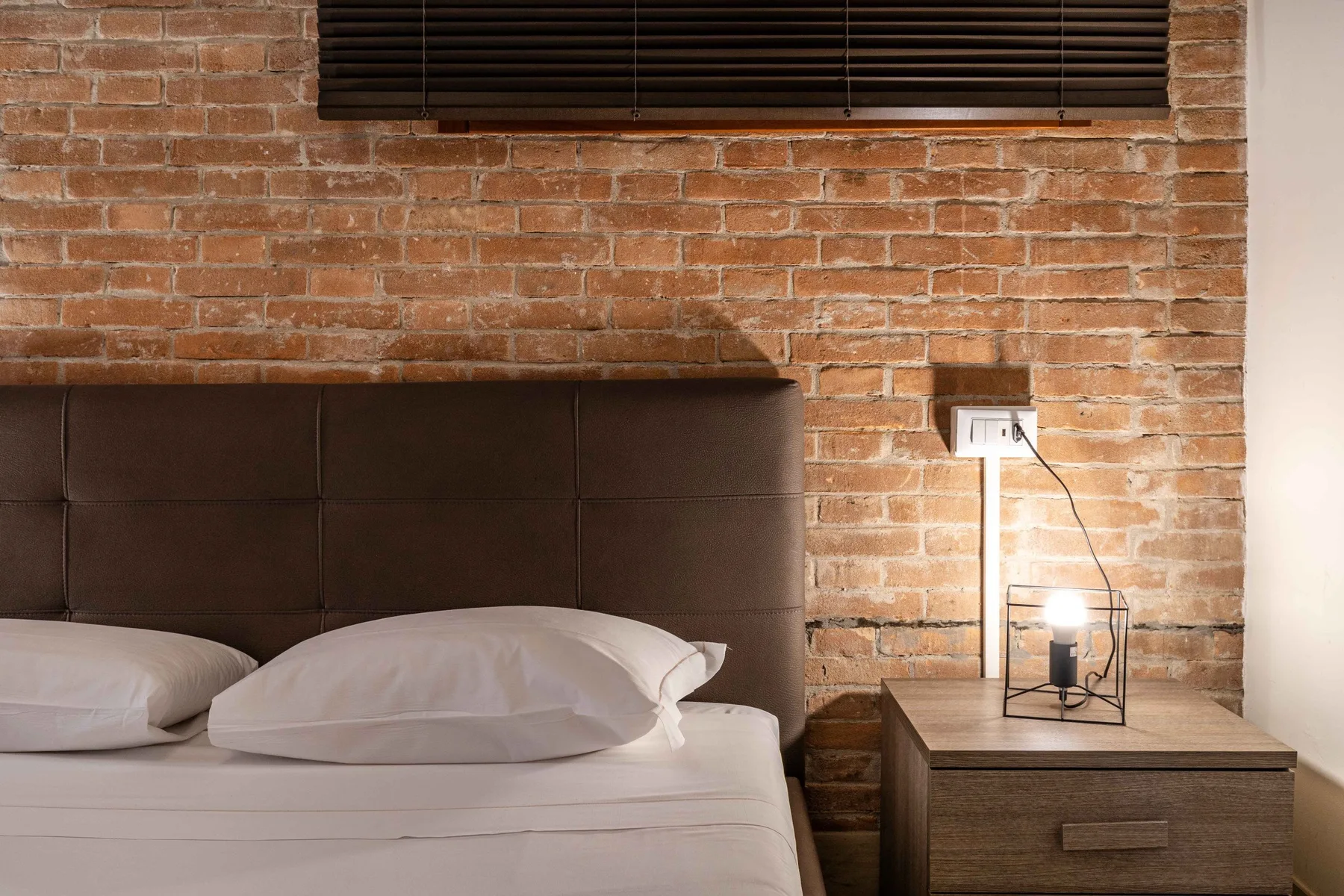 Un dormitorio con una cama junto a una mesita de madera y una pared de ladrillo visto.