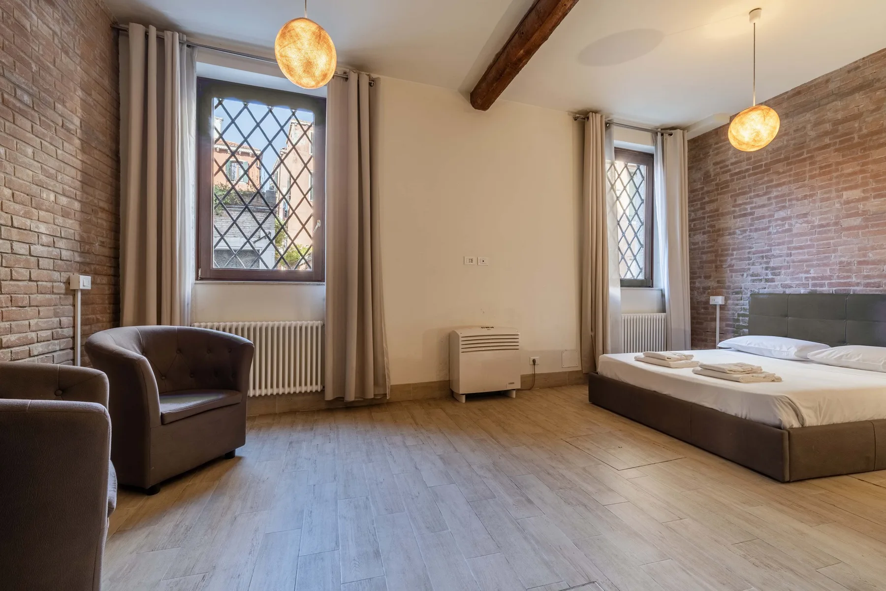 Un dormitorio con una cama, sillones y ventanas altas junto a paredes de ladrillo visto.
