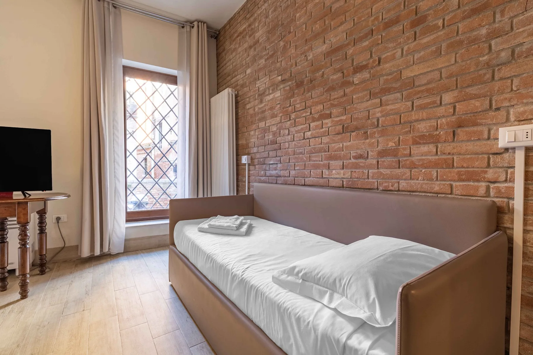 A bedroom with a neatly made bed beside a brick wall and tall window.