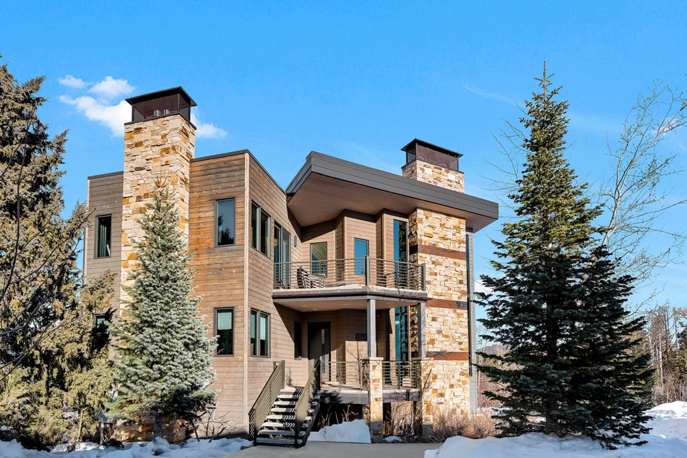 A modern wood-sided house with stone chimneys stands among evergreen trees.