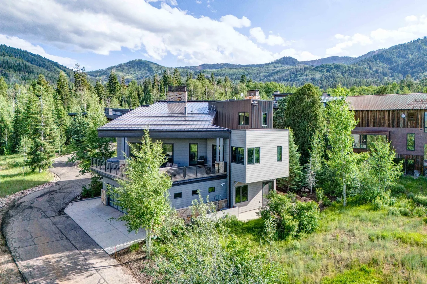 A modern house sits among trees, with forested hills in the background.