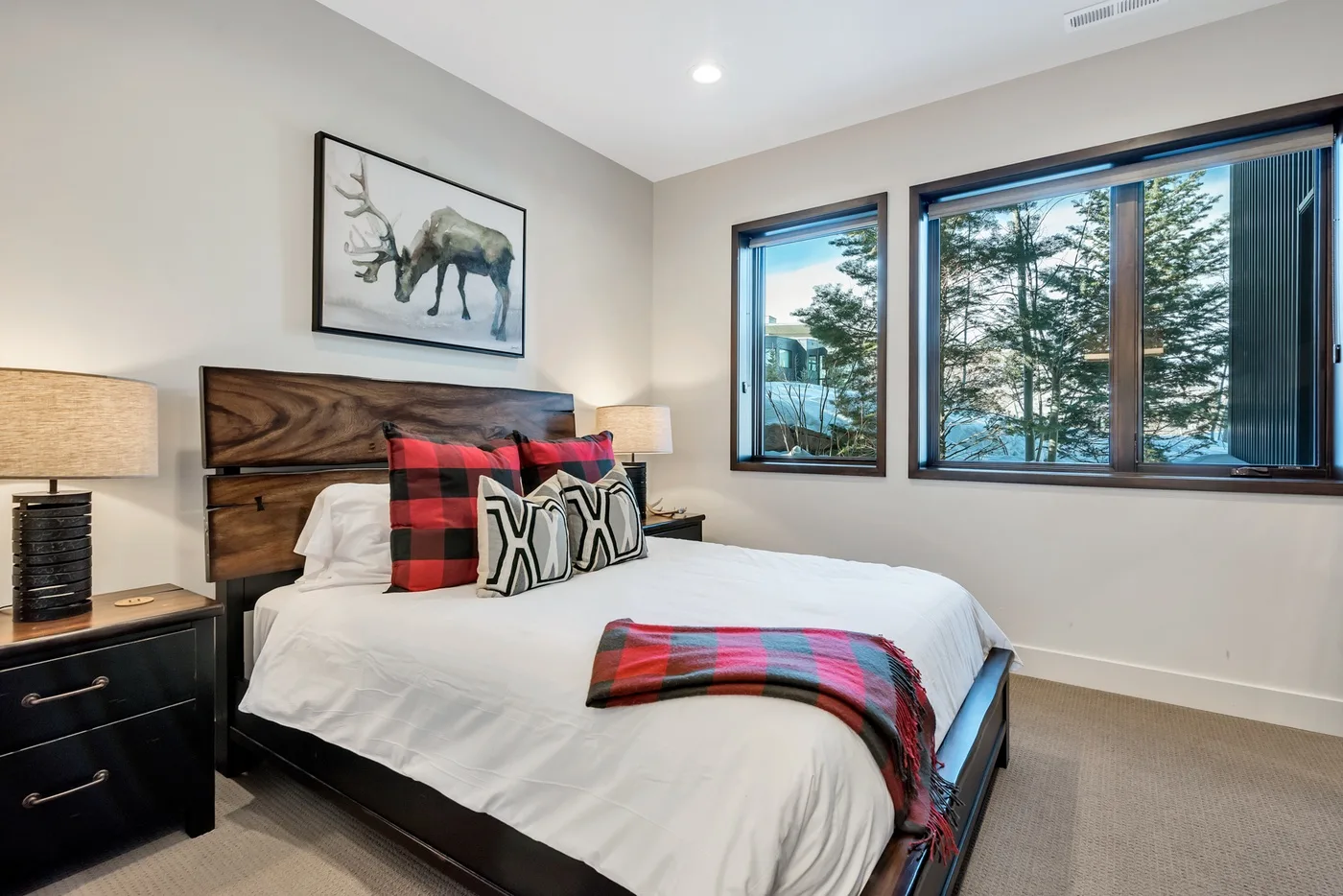 A bedroom with a bed dressed in white and plaid accents beside large windows.