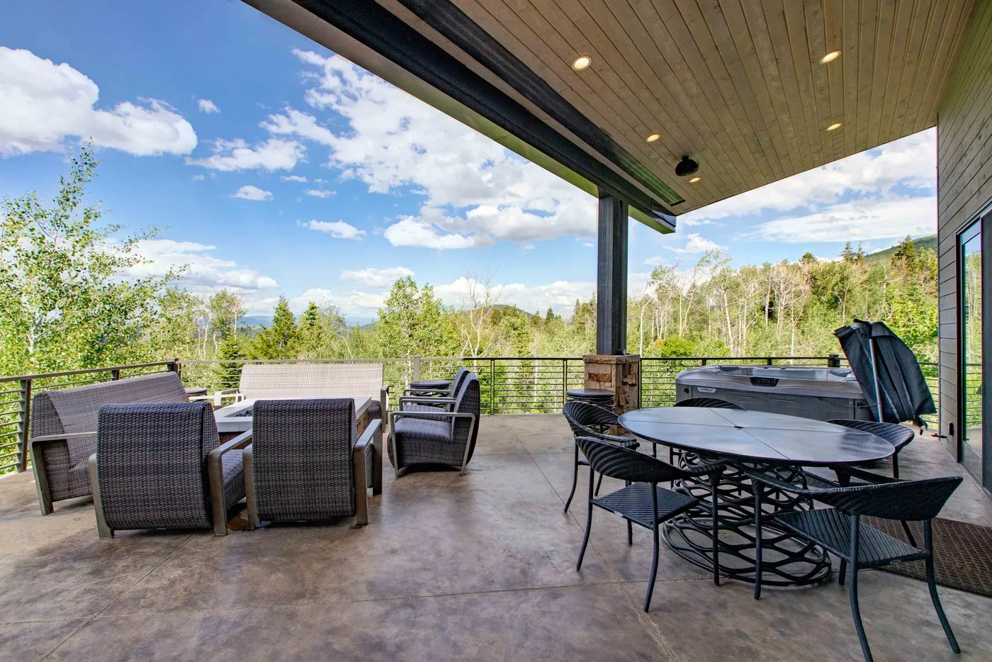 A covered outdoor patio has seating, a dining table, and a hot tub overlooking trees.
