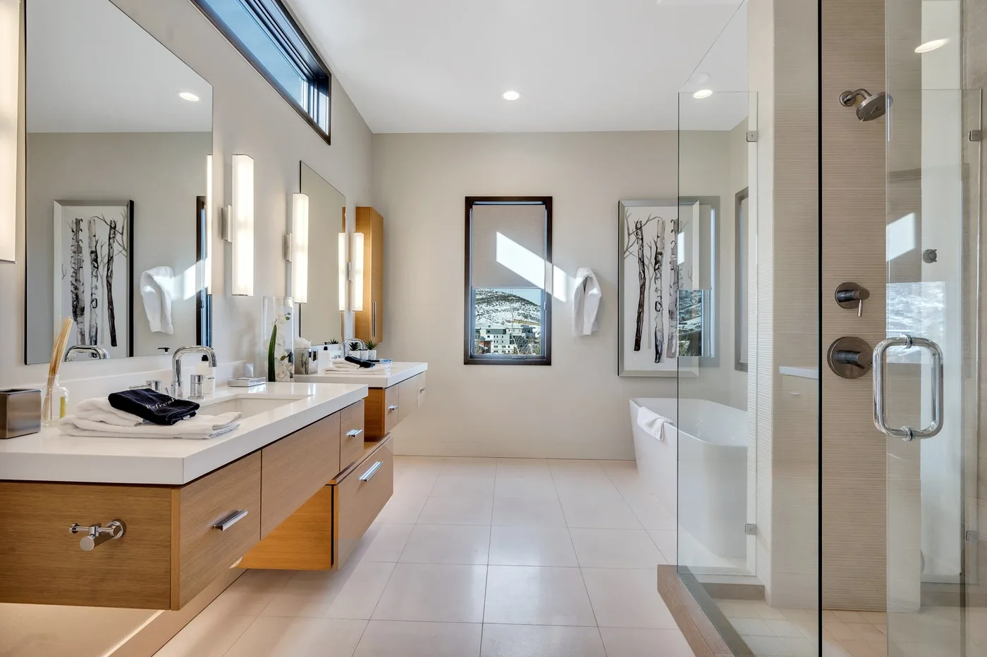 A bright bathroom has a long vanity with sinks and a glass shower.
