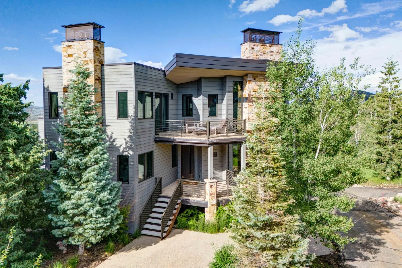 A modern multi-story house with stone chimneys and a wooden deck among trees.