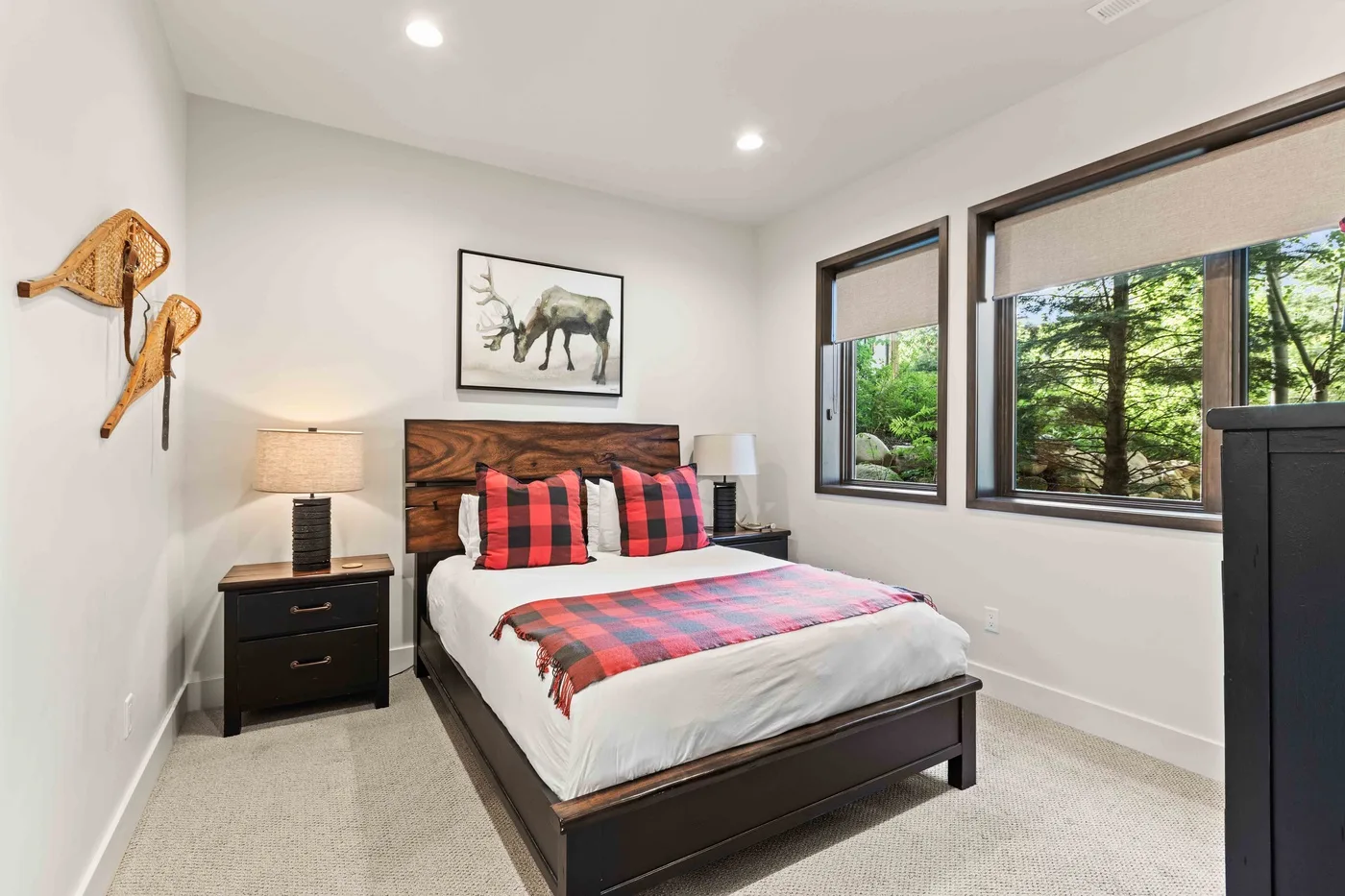 A bedroom with a bed dressed in red-and-black plaid bedding and windows overlooking trees.