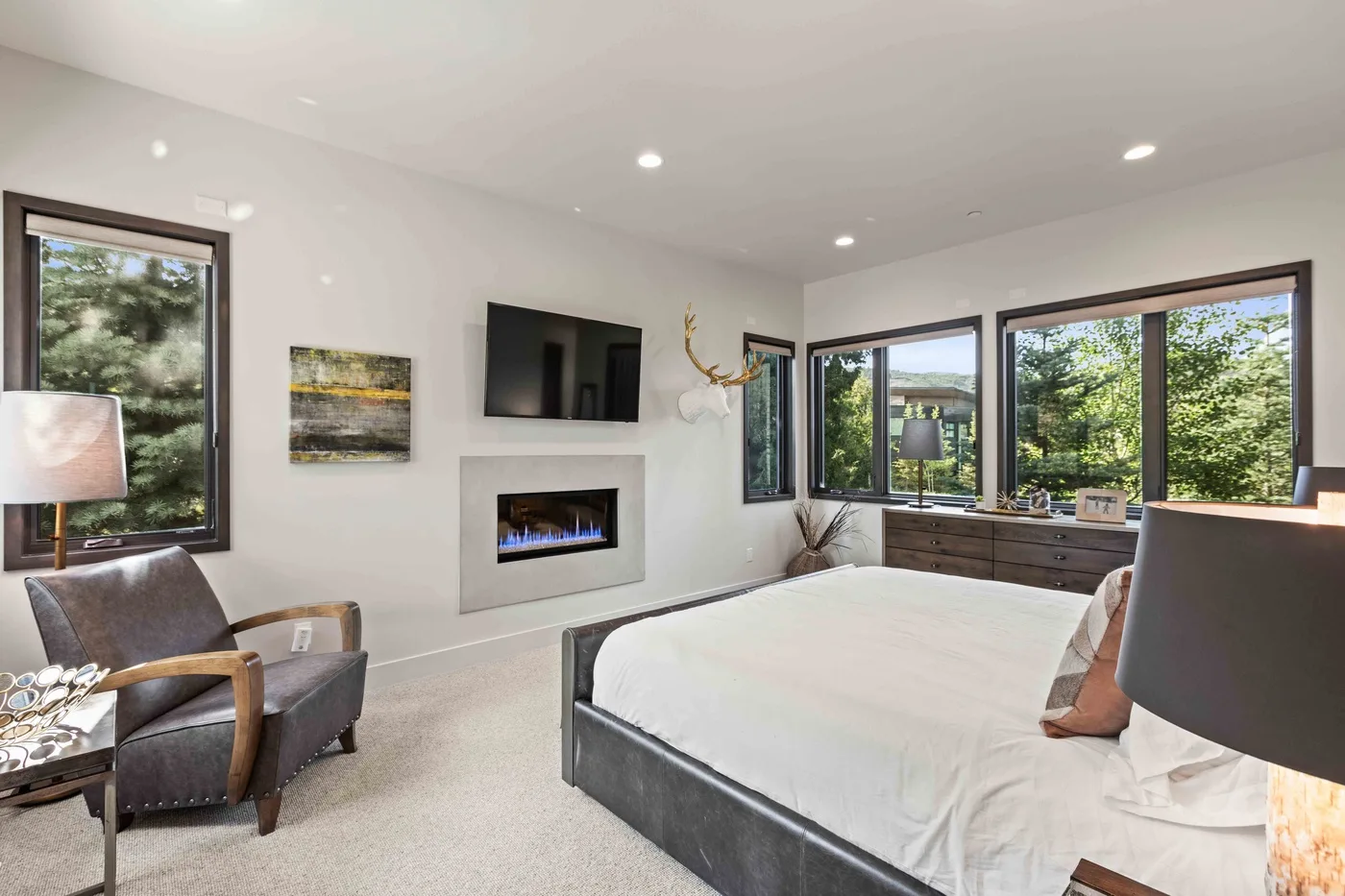A bright bedroom with a bed, wall-mounted television, and large windows overlooking trees.