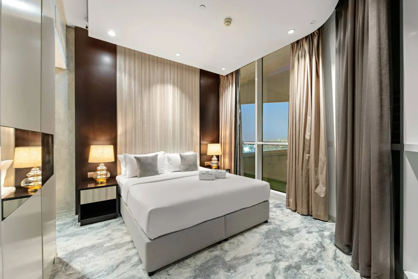 A bedroom with a neatly made bed beside floor-to-ceiling windows and curtains.