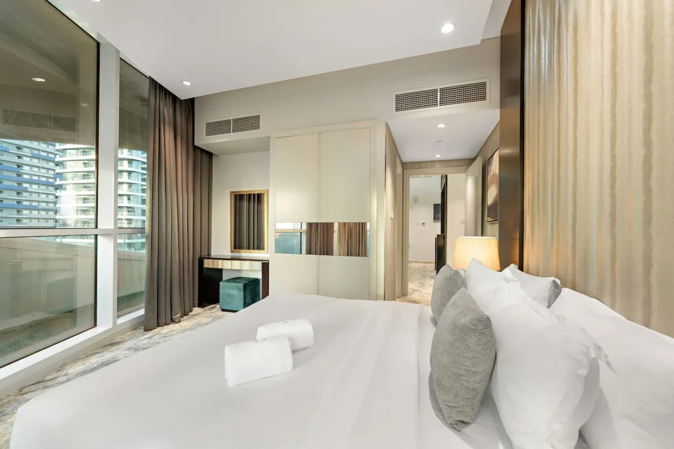 A bedroom with a neatly made bed and large windows overlooking nearby buildings.