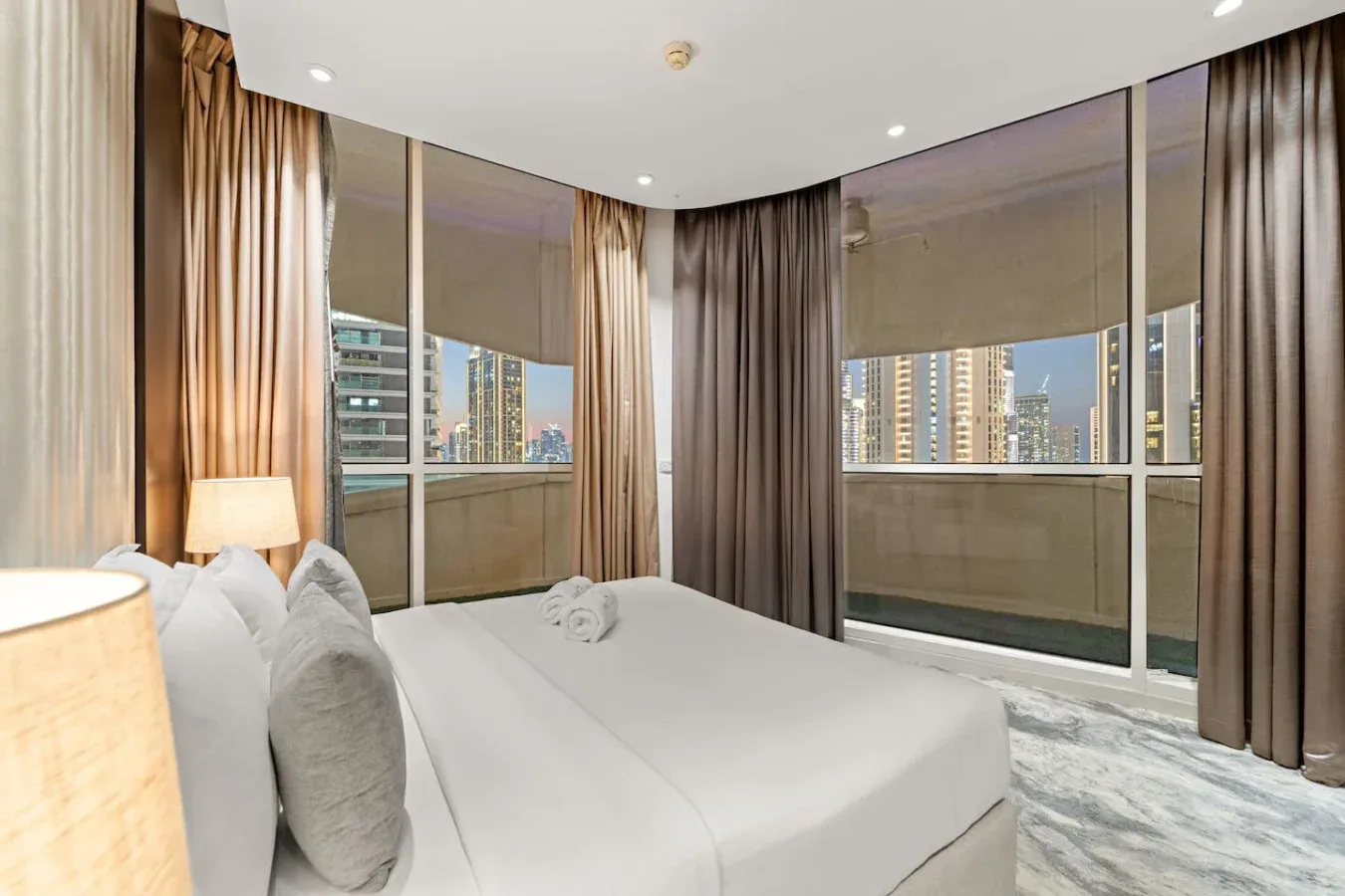 A bedroom with a bed beside floor-to-ceiling windows overlooking nearby buildings.