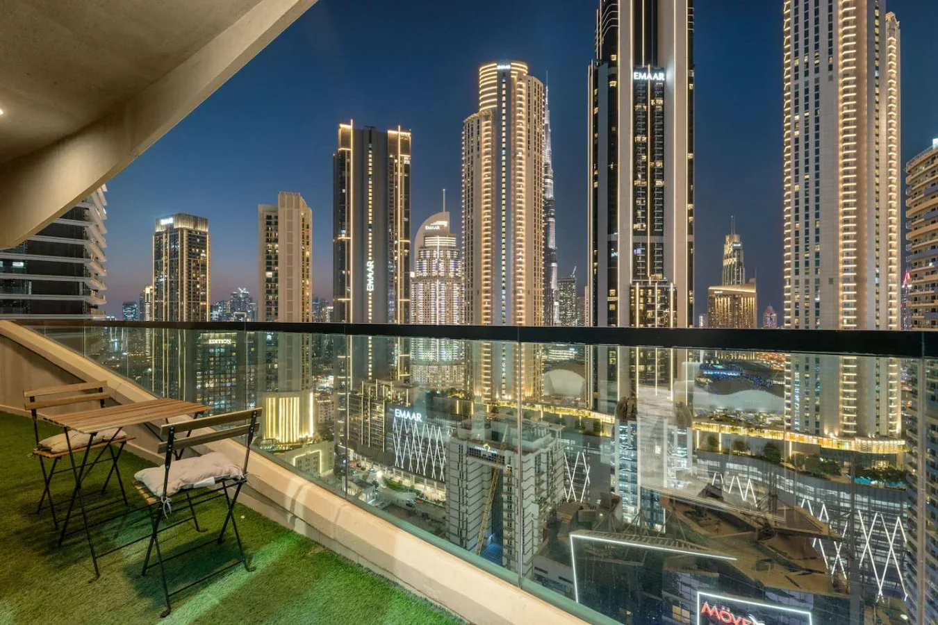 A balcony overlooks illuminated high-rise buildings at night, with outdoor seating beside a glass railing.