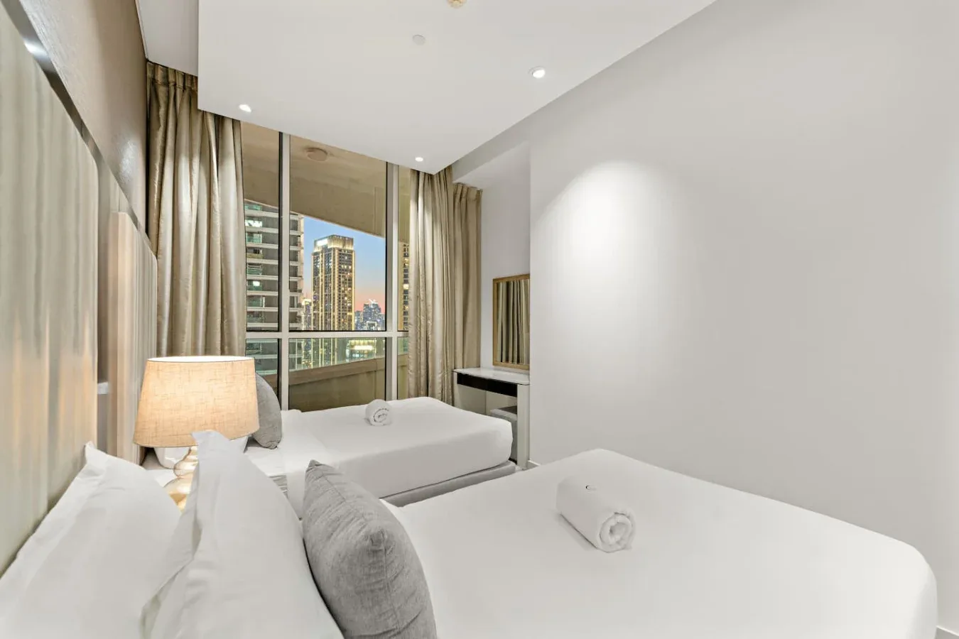 A bright bedroom with two beds, a large window, and city buildings outside.