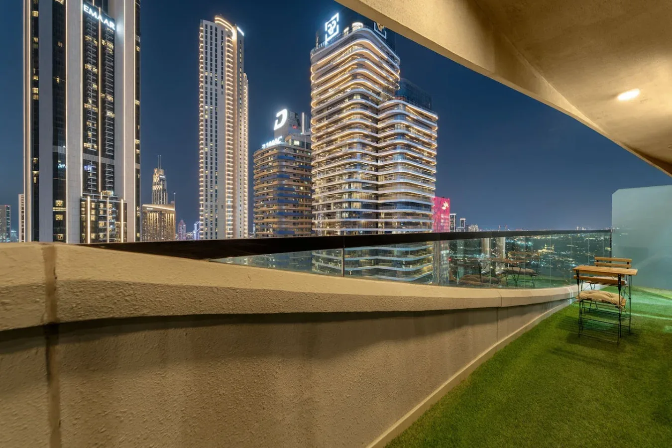 A balcony overlooks illuminated high-rise buildings at night, with a glass railing and green flooring.