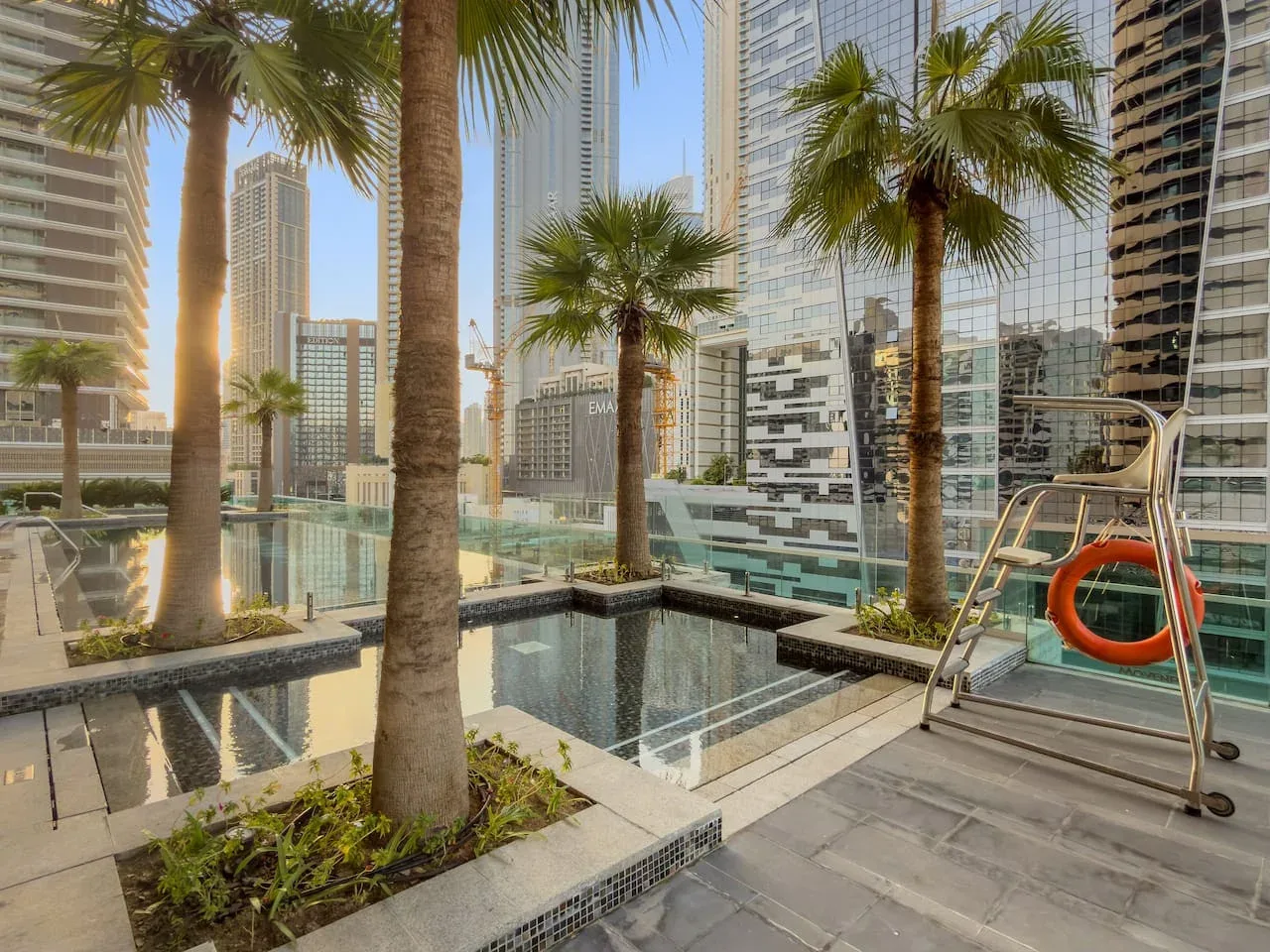 An outdoor swimming pool surrounded by palm trees and modern high-rise buildings.