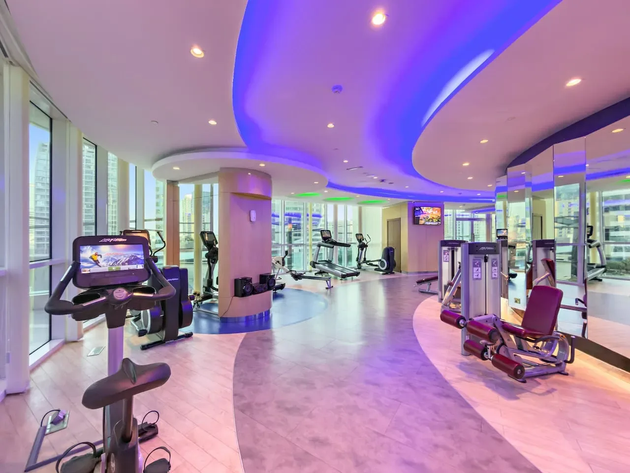 A modern gym with exercise machines, large windows, and blue ceiling lighting.