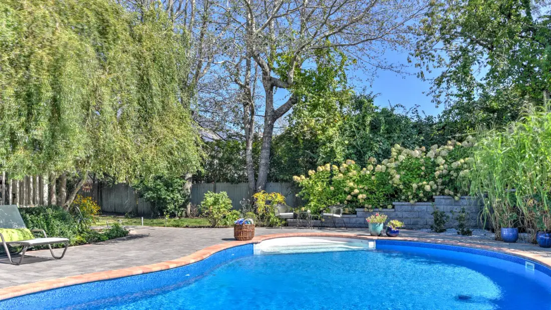 A backyard swimming pool is surrounded by trees, flowering shrubs, and patio seating.