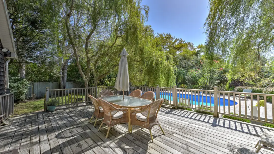 A wooden deck with outdoor seating and a closed umbrella overlooks a swimming pool.