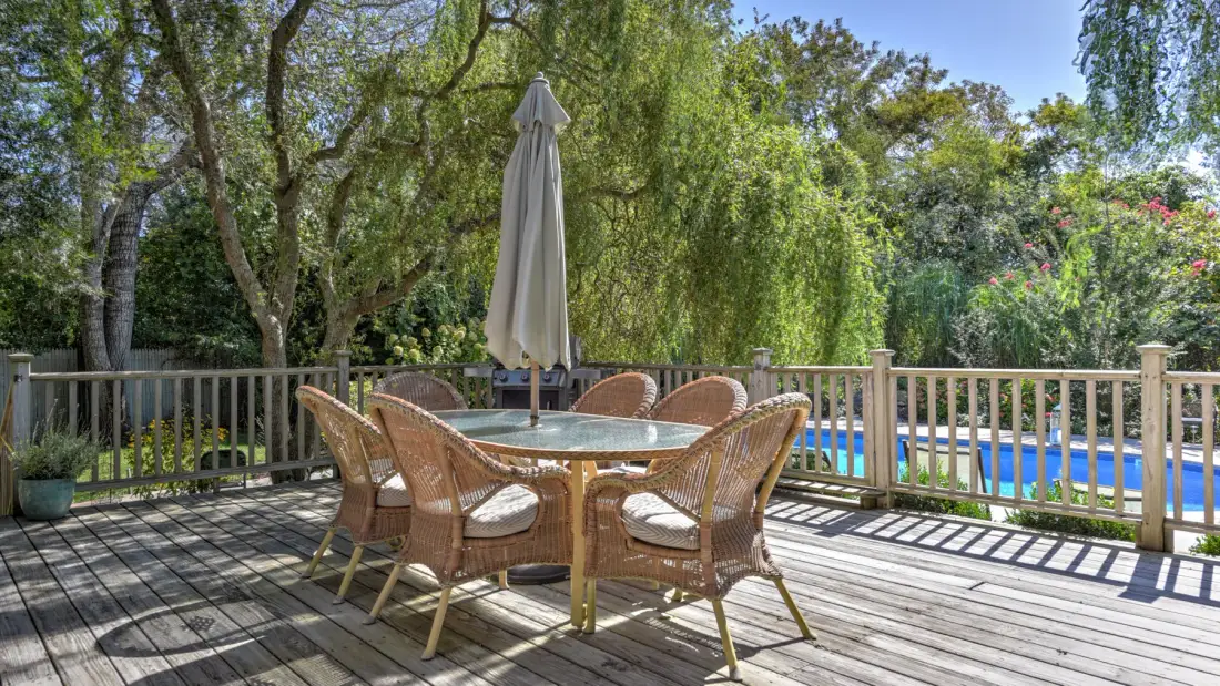 A wooden deck has an outdoor dining table, wicker seating, and a closed umbrella.