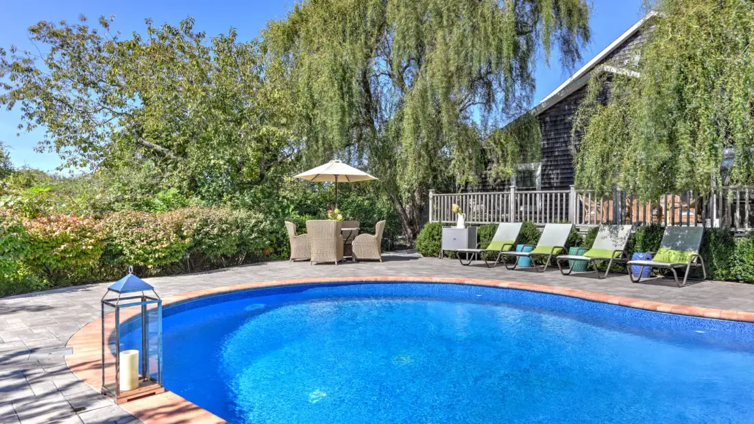 A backyard pool bordered by outdoor seating, trees, and a house.