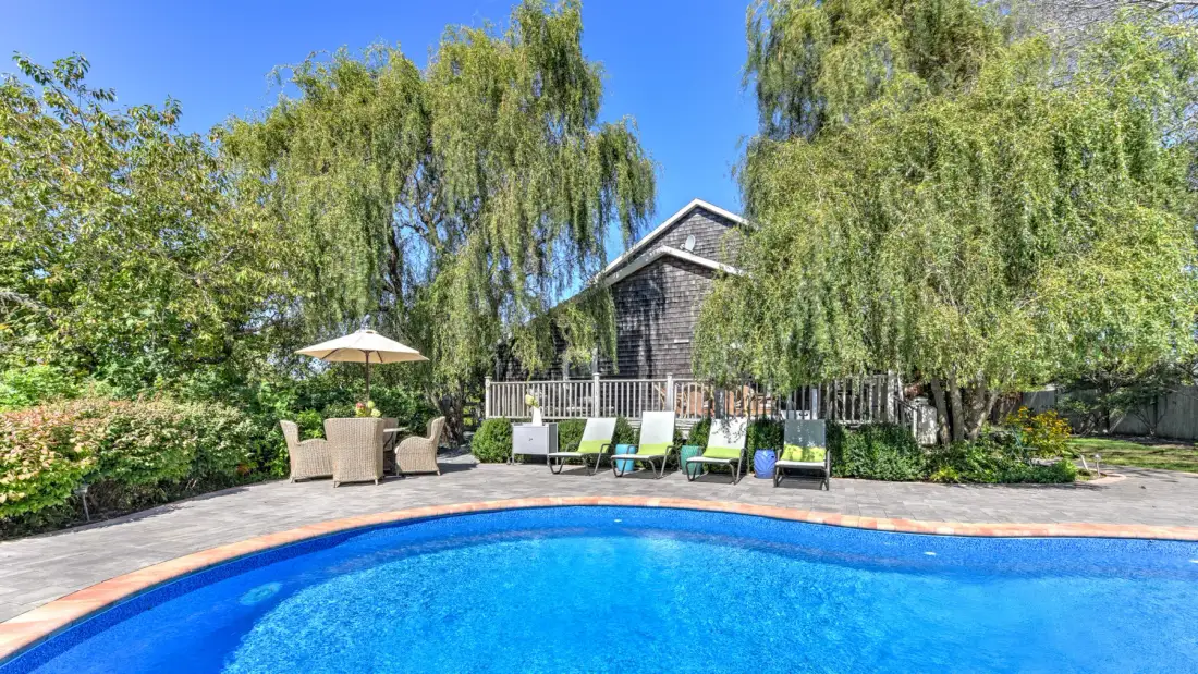 An outdoor swimming pool sits beside a deck with lounge seating and trees.