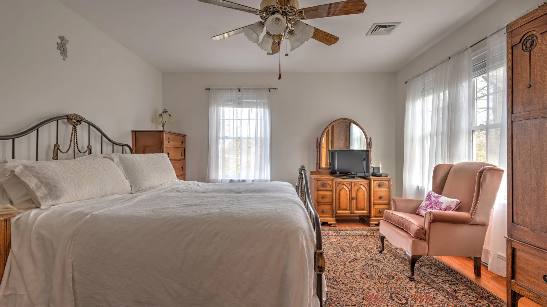A bedroom with a large bed, dresser, and armchair beside bright windows.