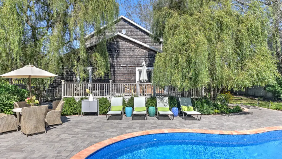 An outdoor pool beside a paved patio with lounge seating and trees.