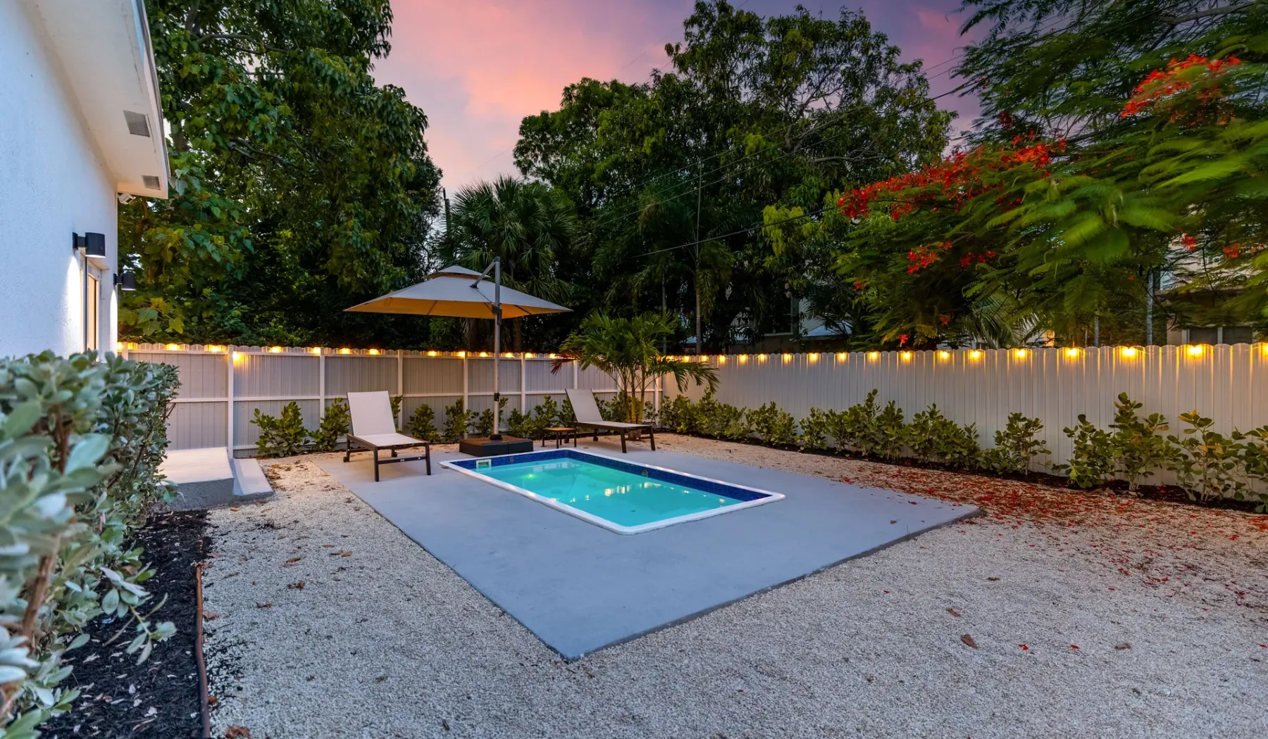 Una piscina al aire libre está en un jardín junto a una cerca iluminada.