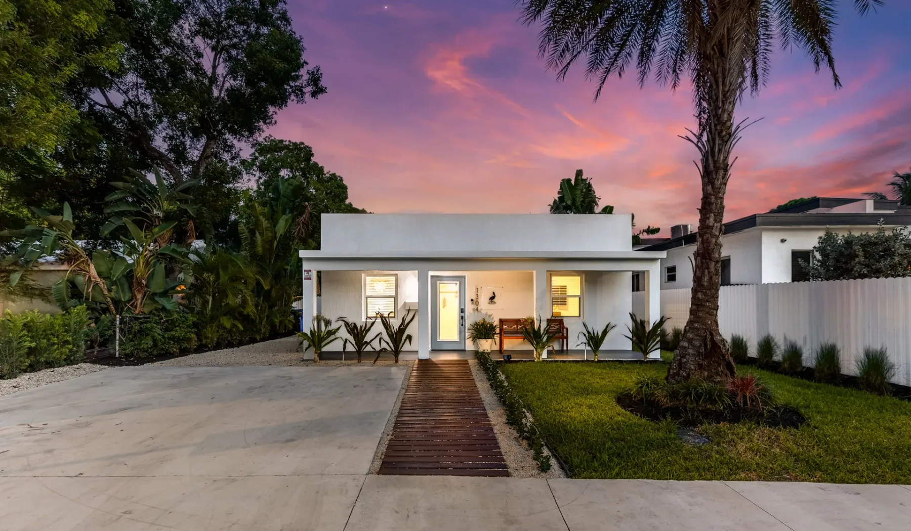 Una casa blanca moderna está junto a una entrada pavimentada bajo un cielo de atardecer colorido.