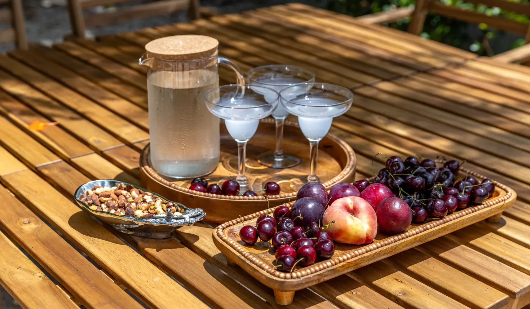 Una mesa de madera al aire libre sostiene fruta, aperitivos, una jarra y copas.
