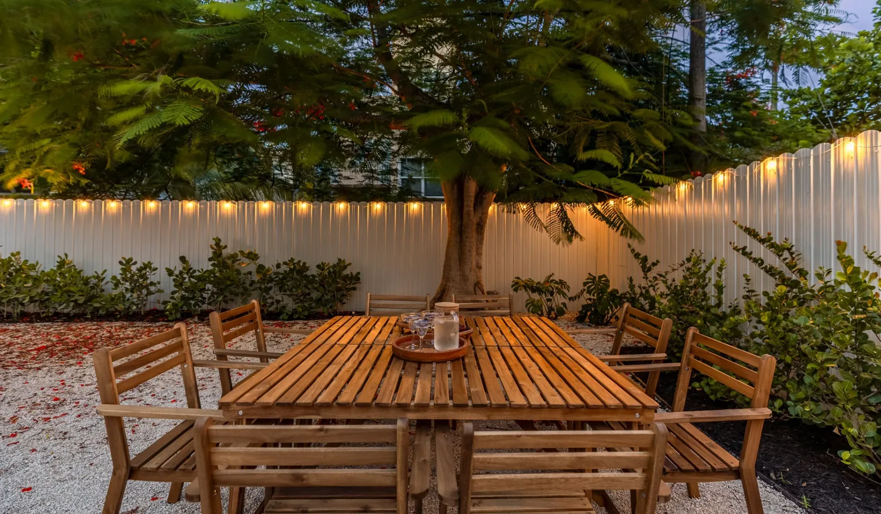 Una mesa de comedor de madera y asientos están bajo un árbol en un patio cercado.