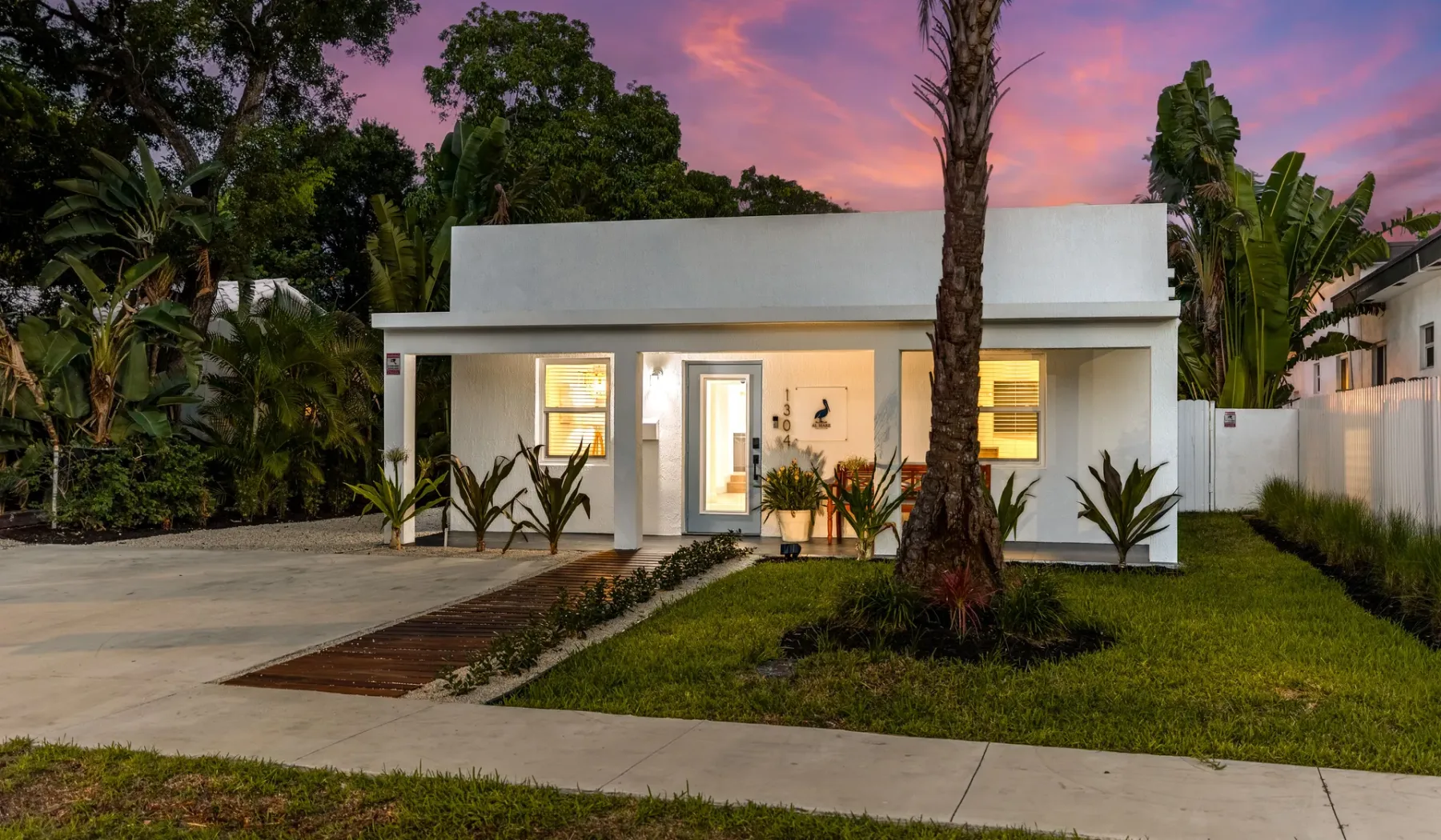 Una casa blanca moderna se encuentra junto a una entrada pavimentada y un jardín al atardecer.
