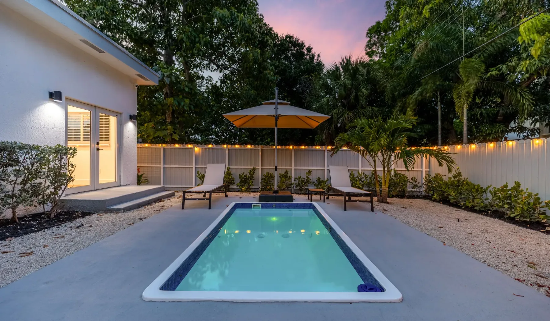 Una piscina al aire libre está junto a un patio con una sombrilla y vegetación alrededor.