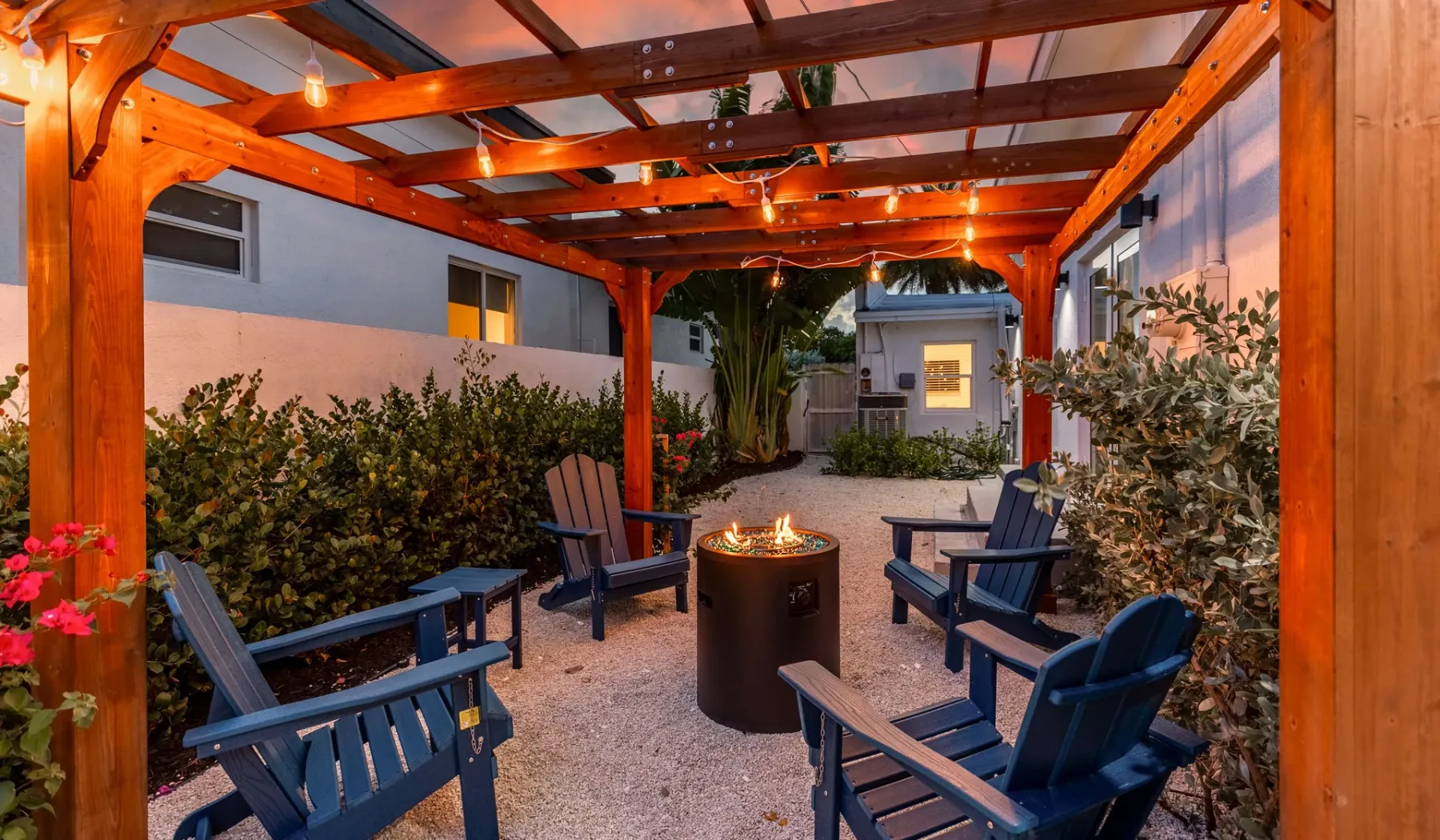 Un patio exterior con asientos alrededor de una fogata, bajo una pérgola de madera.