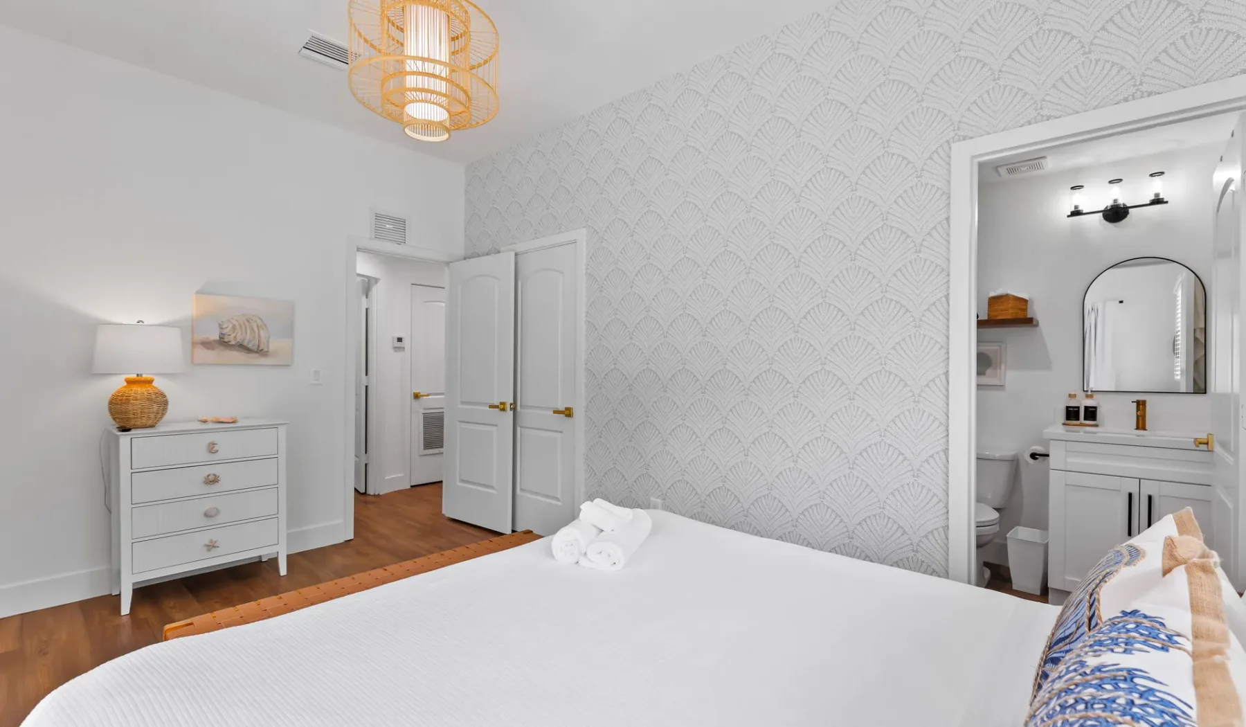 Un dormitorio con una cama blanca, papel tapiz estampado y una entrada a un baño.
