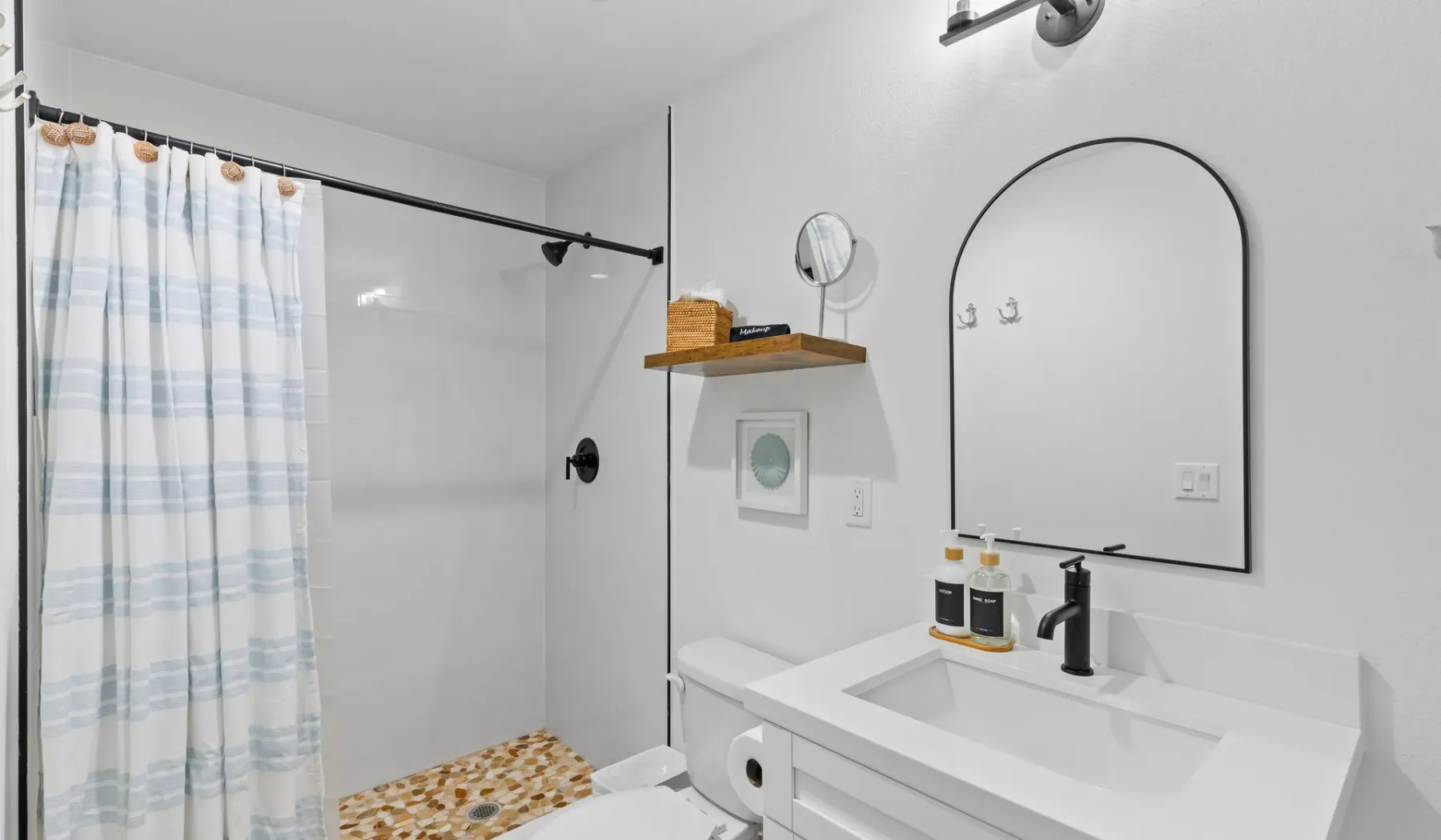 Un baño luminoso con ducha, cortina a rayas y lavabo bajo un espejo arqueado.