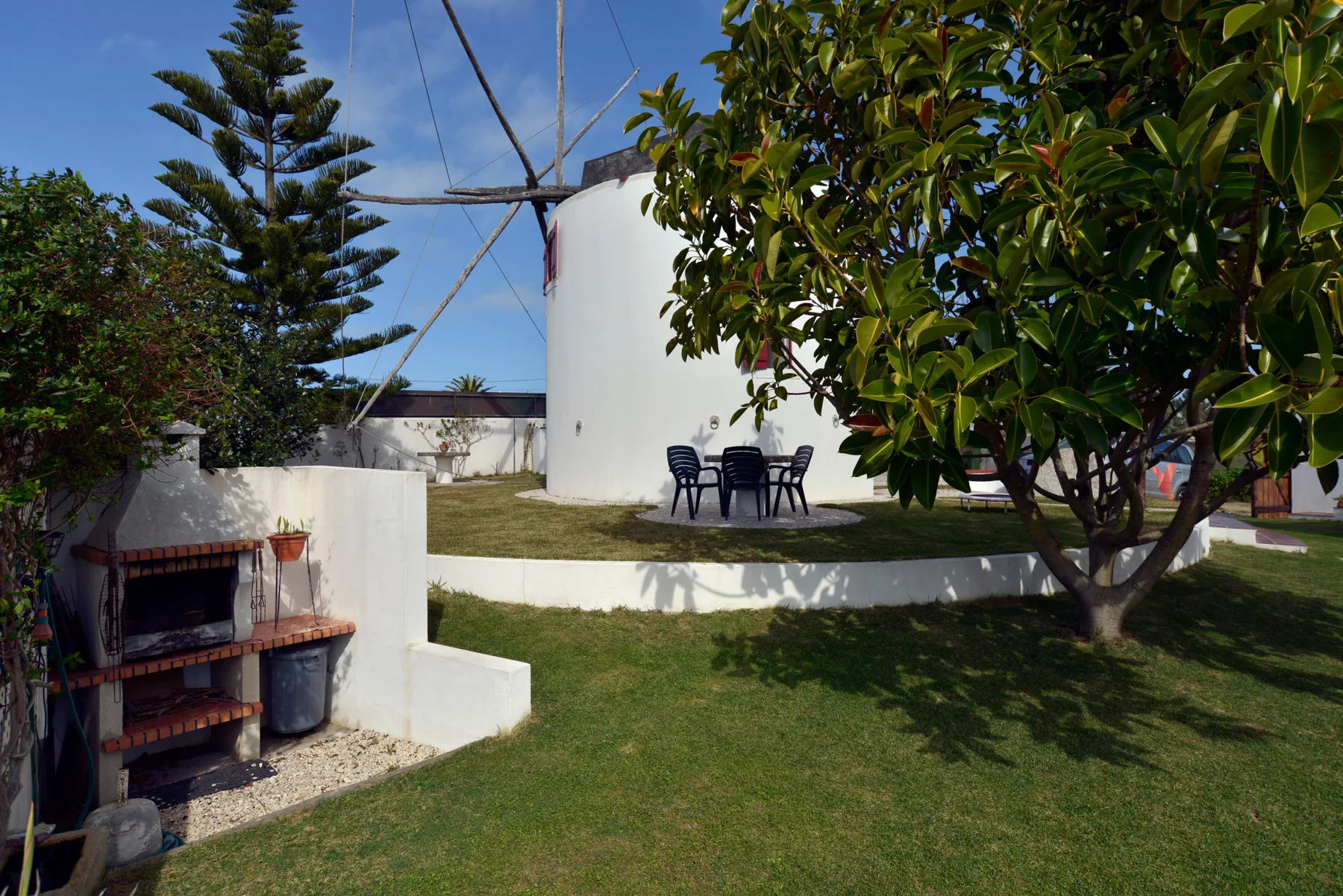 Un patio exterior con césped junto a un molino blanco y asientos bajo un árbol frondoso.