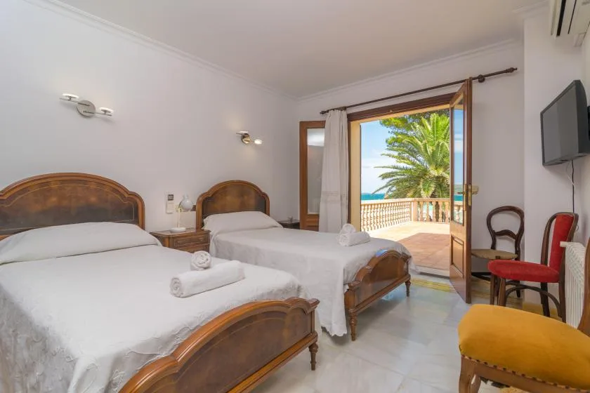 Un dormitorio con dos camas y una puerta abierta al balcón, frente a palmeras.