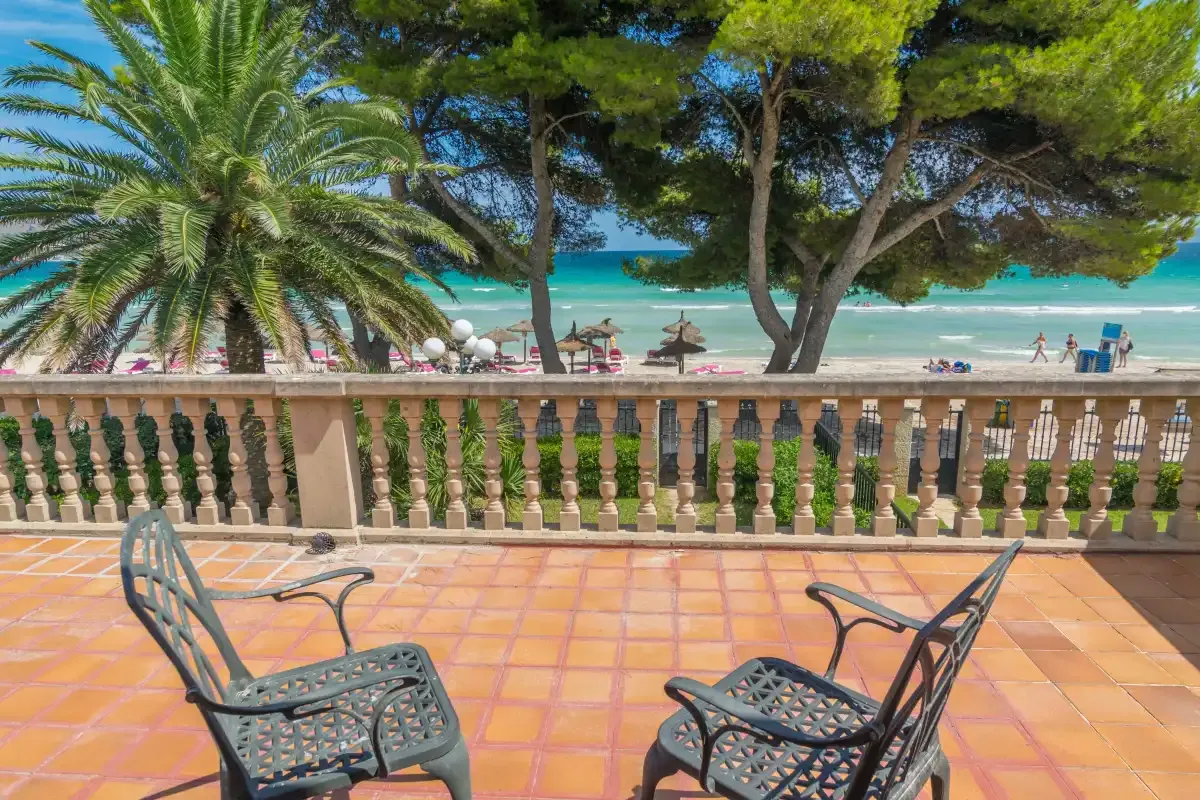 Una terraza con suelo de baldosas y asientos metálicos da a palmeras y una playa de arena.