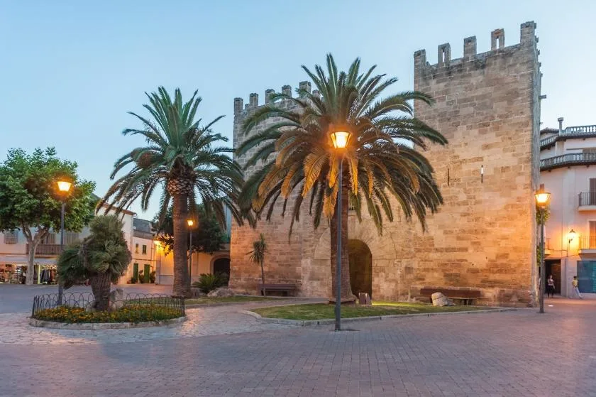 Una muralla y torre de piedra junto a palmeras bordean una plaza pavimentada.