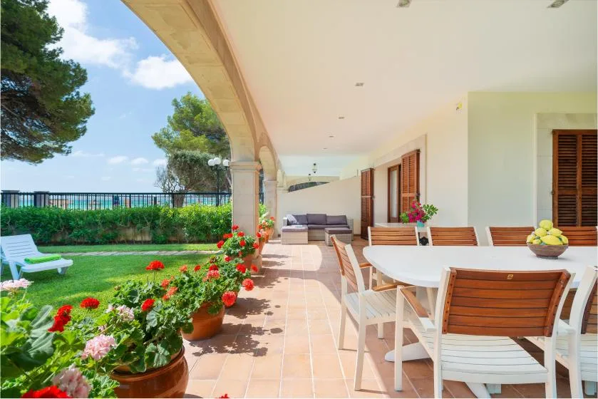 Un patio cubierto tiene una mesa de comedor junto a un jardín y césped verde.